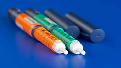 Two insulin pens, one orange and one green, lie on a blue surface; two dark blue pen caps are in the background.