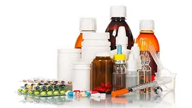 Assorted medications: pill bottles, liquid medicine, blister packs, and a syringe, on a reflective surface against a white background.
